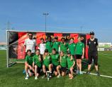 Eine Gruppe junger Frauen und Mädchen in Sportuniformen posiert vor einem Hintergrundbanner, wahrscheinlich im Rahmen eines Turniers auf einem grünen