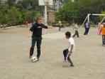 Ein Mann und zwei Jungen spielen Fußball auf einem Schotterplatz, während weitere Spieler im Hintergrund sichtbar sind.