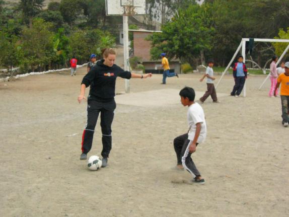 Ein Mann und zwei Jungen spielen Fußball auf einem Schotterplatz, während weitere Spieler im Hintergrund sichtbar sind.