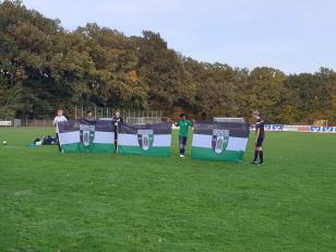 Eine Gruppe von Menschen hält auf einem grünen Sportfeld verschiedene Vereinsflaggen vor einem Hintergrund aus Bäumen.