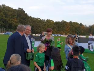Eine Gruppe von Menschen, darunter Erwachsene und Kinder, steht auf einem grünen Sportplatz.