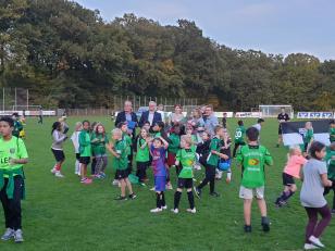 Eine große Gruppe von Kindern in Sportuniformen steht auf einem grünen Fußballfeld vor einem bewaldeten Hintergrund.