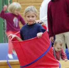 Mehrere Kinder stehen um einen großen roten Behälter, während im Hintergrund erwachsene Personen zu sehen sind.