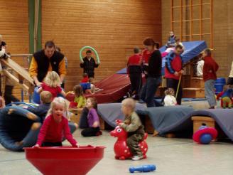 Viele spielende Kinder und Erwachsene in einer großen Turnhalle mit Spielgeräten und Matten.