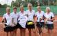 Gruppe von acht Frauen in Sportkleidung posieren vor einem Tennisplatz.