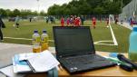 Ein Laptop und Dokumente auf einem Tisch mit Blick auf einen belebten Sportplatz im Hintergrund.