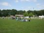 Ein großer, grüner Sportplatz mit Menschen und Bäumen im Hintergrund unter blauem Himmel.