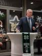Ein Mann in Anzug präsentiert goldene Trophäen bei einem Event vor einem Tisch mit Vereinslogo.