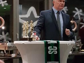 Ein Mann in Anzug präsentiert goldene Trophäen bei einem Event vor einem Tisch mit Vereinslogo.