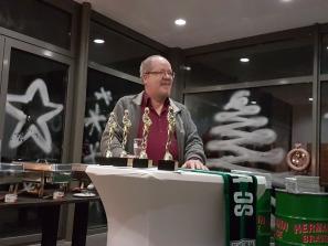 Ein Mann steht hinter einem Podium, das mit zahlreichen goldenen Trophäen und Bannern des SC V geschmückt ist.