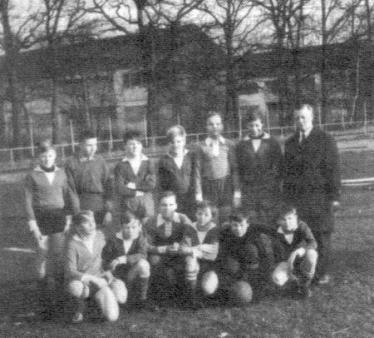 Eine große Gruppe von Männern und jungen Männern posiert draußen in einer lockeren Formation vor einem Gebäude.