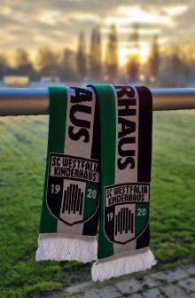 Drei sportliche Schals in Grün, Schwarz und Weiß liegen auf einem Rasen, mit einem unscharfen Sonnenuntergangshintergrund.