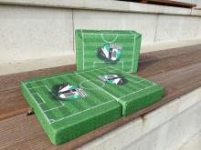Mehrere grüne Kunstrasen-Matten, die einen Miniaturfußballplatz darstellen, liegen auf einer Holzbank.