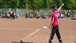 Ein Junge in einem roten Fußballtrikot steht auf einem Sportplatz, während im Hintergrund viele Menschen und Bäume zu sehen sind.