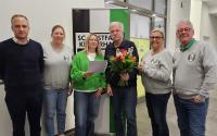 Eine Gruppe von sieben Personen, bestehend aus Männern und Frauen, posiert in einem Innenraum vor einem Informationsdisplay.